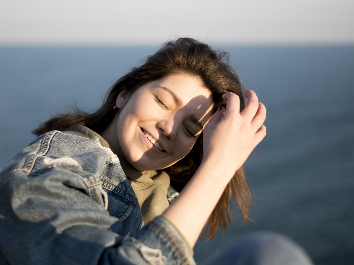 A woman smiling with her eyes closed in warm sunlight by the sea