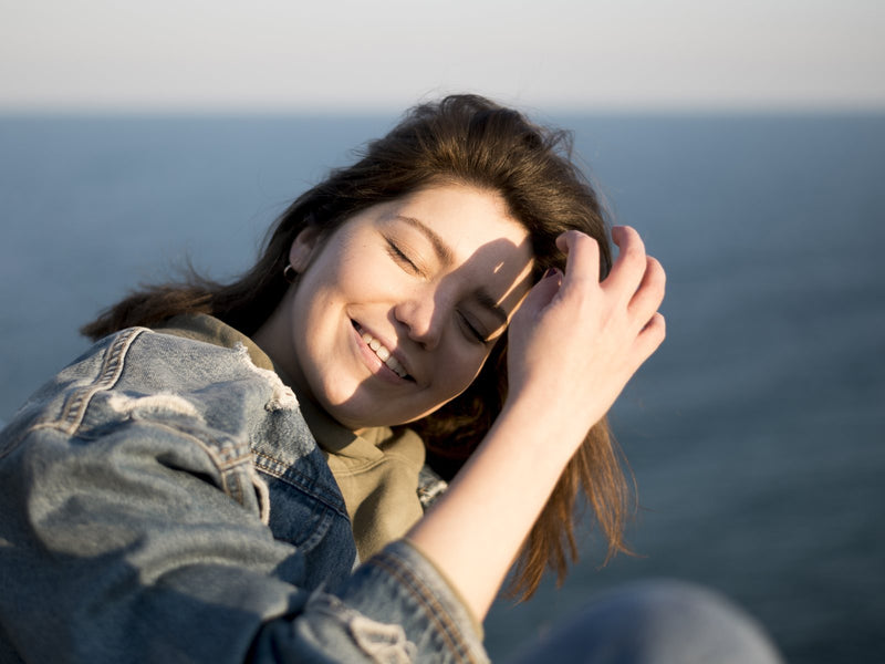 A woman smiling with her eyes closed in warm sunlight by the sea