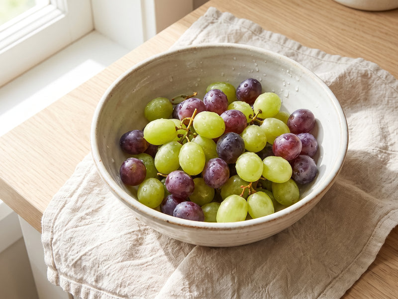 A bowl of red and green grapes on linen by a sunny window